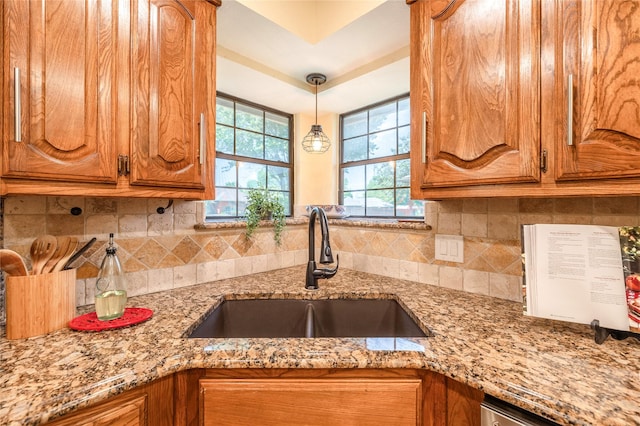kitchen featuring backsplash, a sink, light stone countertops, brown cabinetry, and hanging light fixtures