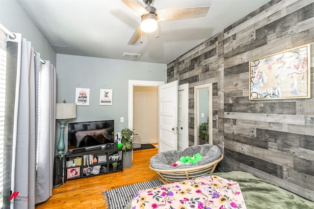 living area featuring a ceiling fan, an accent wall, wood finished floors, wooden walls, and visible vents
