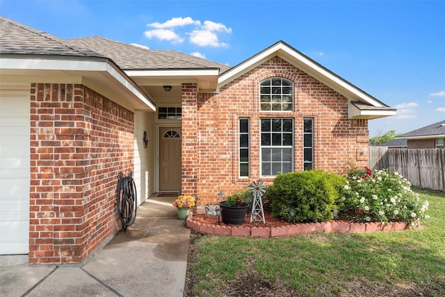 doorway to property with fence, roof with shingles, an attached garage, a lawn, and brick siding