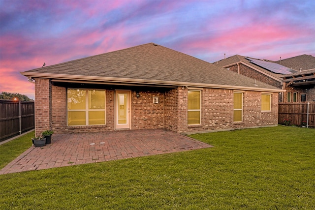 back of house at dusk featuring a patio, roof with shingles, a lawn, and a fenced backyard