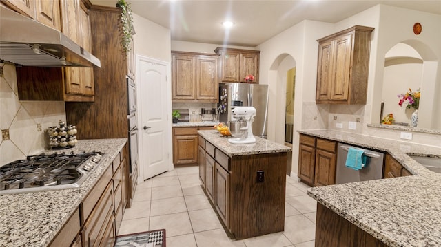 kitchen featuring light tile patterned floors, appliances with stainless steel finishes, backsplash, under cabinet range hood, and light stone countertops