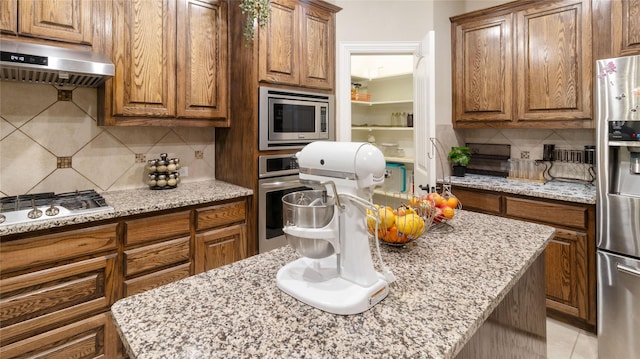 kitchen featuring light stone counters, brown cabinetry, appliances with stainless steel finishes, backsplash, and under cabinet range hood