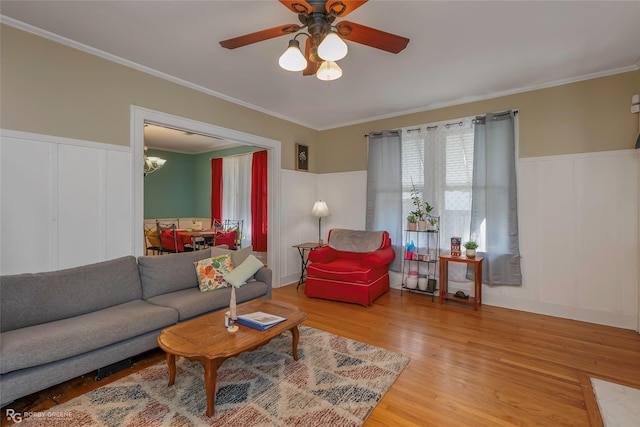 living room featuring wainscoting, ceiling fan with notable chandelier, wood finished floors, a decorative wall, and ornamental molding