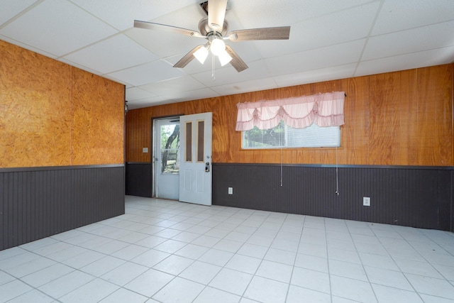 empty room featuring wooden walls, light tile patterned floors, ceiling fan, a wainscoted wall, and a paneled ceiling