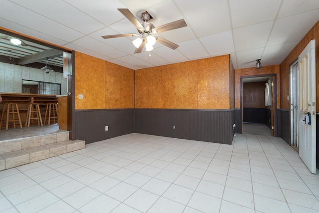 empty room featuring wainscoting, wooden walls, ceiling fan, and a drop ceiling