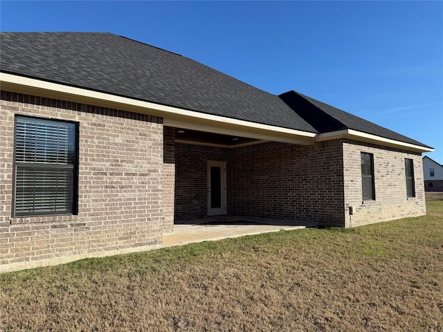 rear view of property featuring brick siding, a lawn, a patio, and roof with shingles