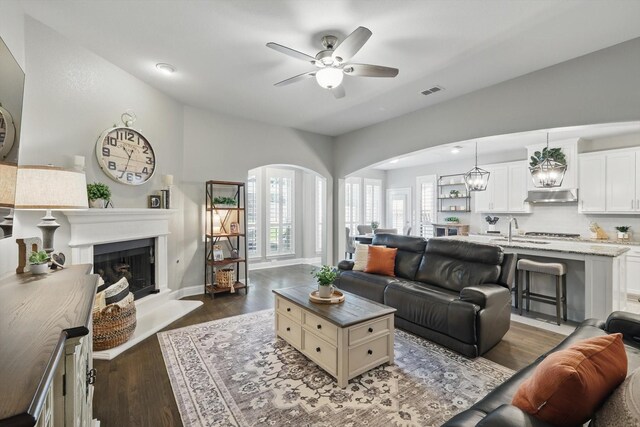 living area featuring ceiling fan, arched walkways, a fireplace with raised hearth, dark wood-type flooring, and visible vents