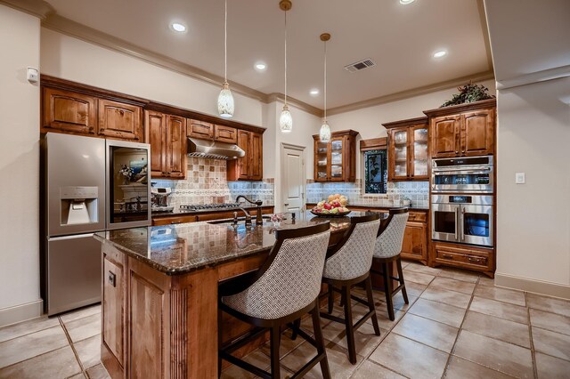 kitchen featuring under cabinet range hood, ornamental molding, a center island with sink, tasteful backsplash, and appliances with stainless steel finishes