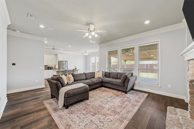 living area featuring dark wood finished floors, baseboards, ceiling fan, crown molding, and a stone fireplace