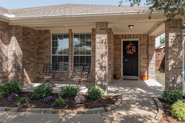 view of exterior entry featuring a shingled roof, covered porch, and brick siding