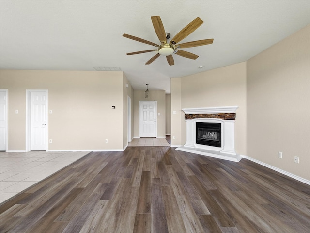 unfurnished living room with wood finished floors, a fireplace with raised hearth, a ceiling fan, and baseboards
