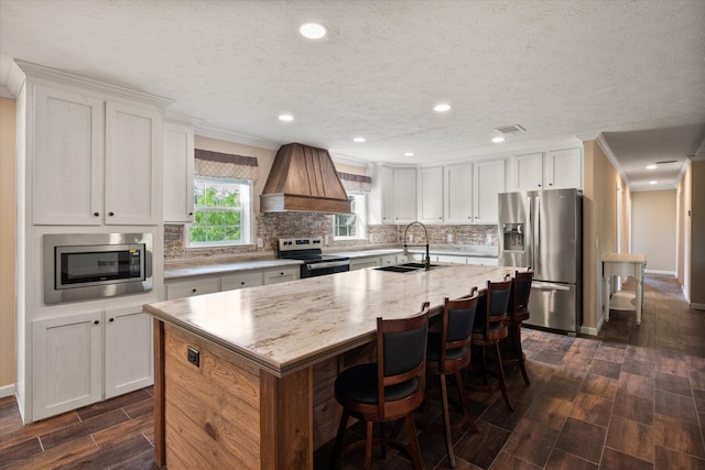 kitchen featuring wood tiled floor, an island with sink, stainless steel appliances, visible vents, and custom exhaust hood
