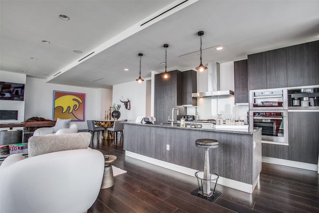 kitchen with wall chimney exhaust hood, a sink, an island with sink, modern cabinets, and dark wood-type flooring