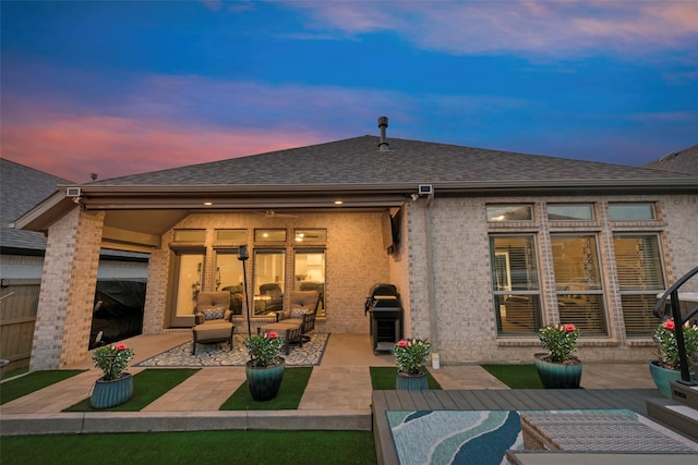 back of property at dusk with brick siding, outdoor lounge area, a patio area, and roof with shingles