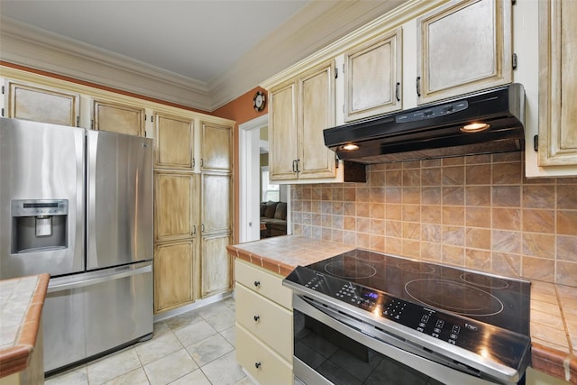 kitchen with tile countertops, ornamental molding, stainless steel appliances, backsplash, and under cabinet range hood