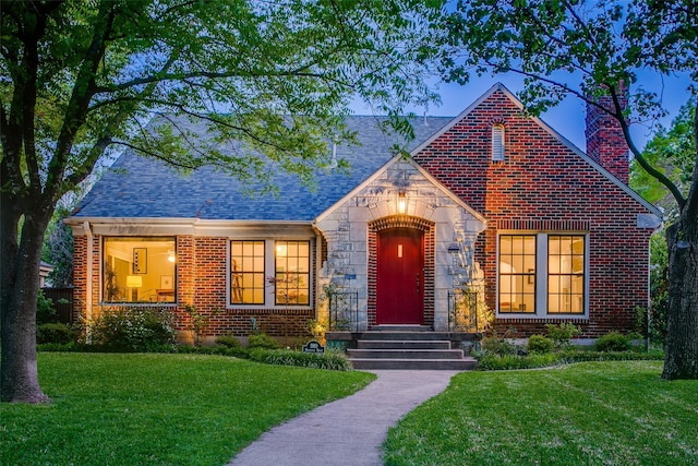 tudor home with roof with shingles, a front lawn, stone siding, a chimney, and brick siding