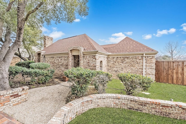 french country home featuring brick siding, roof with shingles, a chimney, and fence