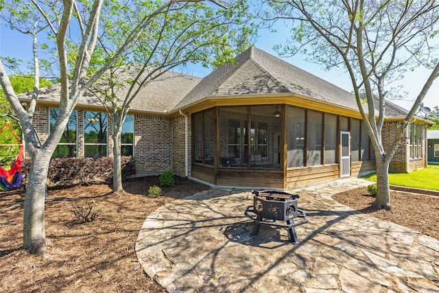 rear view of property featuring brick siding, a patio area, a fire pit, roof with shingles, and a sunroom