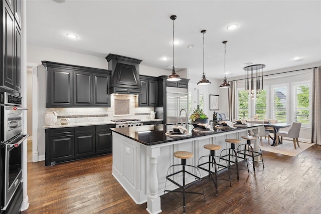 kitchen featuring stainless steel appliances, a center island with sink, tasteful backsplash, a breakfast bar area, and premium range hood