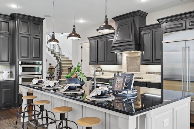 kitchen with stainless steel appliances, premium range hood, a kitchen island with sink, tasteful backsplash, and dark wood-style flooring