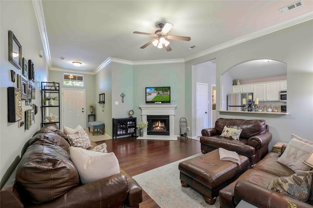 living room featuring a lit fireplace, a ceiling fan, dark wood-style flooring, visible vents, and ornamental molding