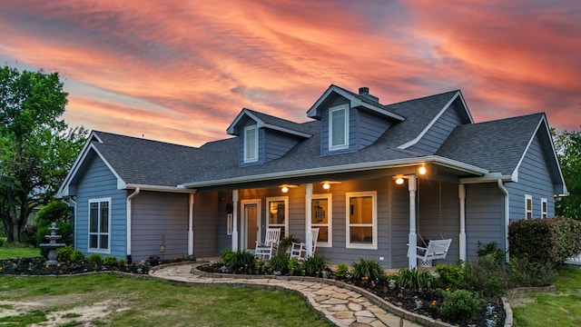 new england style home with a shingled roof, a chimney, covered porch, and a front yard