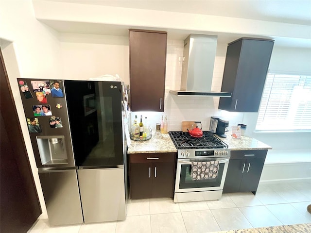 kitchen with wall chimney range hood, decorative backsplash, light tile patterned floors, appliances with stainless steel finishes, and light stone countertops