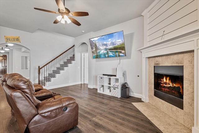 living room with dark wood-style floors, arched walkways, a tiled fireplace, stairs, and ceiling fan