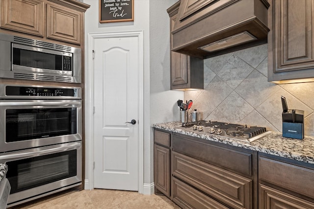 kitchen featuring stainless steel appliances, light stone counters, backsplash, dark brown cabinets, and custom exhaust hood