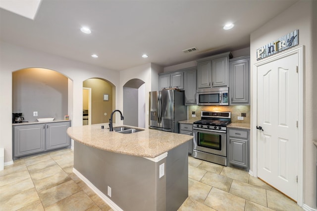 kitchen featuring tasteful backsplash, a sink, a kitchen island with sink, appliances with stainless steel finishes, and gray cabinetry