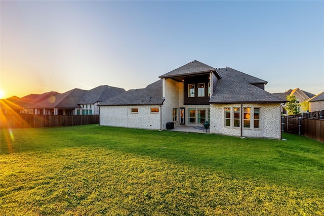 back of property at dusk with a fenced backyard, a yard, a patio, and a shingled roof