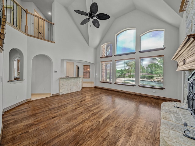 unfurnished living room featuring a fireplace with raised hearth, wood finished floors, a ceiling fan, arched walkways, and high vaulted ceiling