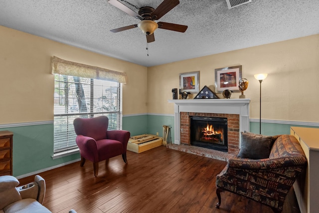 sitting room with a textured ceiling, visible vents, a ceiling fan, wood finished floors, and a brick fireplace