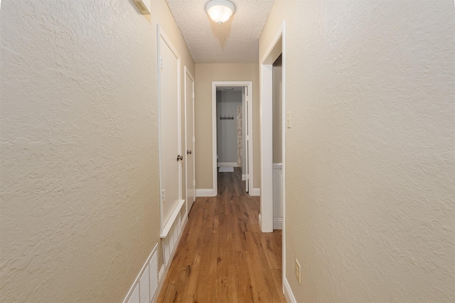 corridor with a textured ceiling, a textured wall, light wood finished floors, and baseboards