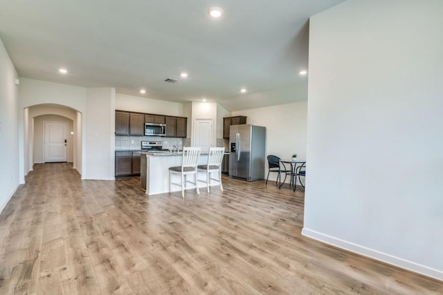 kitchen with stainless steel appliances, arched walkways, light wood-type flooring, dark brown cabinetry, and an island with sink
