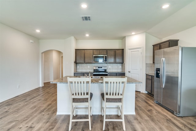 kitchen with dark brown cabinets, visible vents, arched walkways, a center island with sink, and appliances with stainless steel finishes