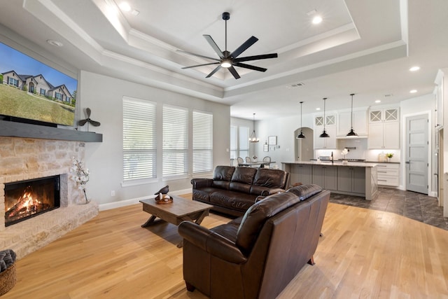 living area with a ceiling fan, light wood-style flooring, a stone fireplace, and a raised ceiling