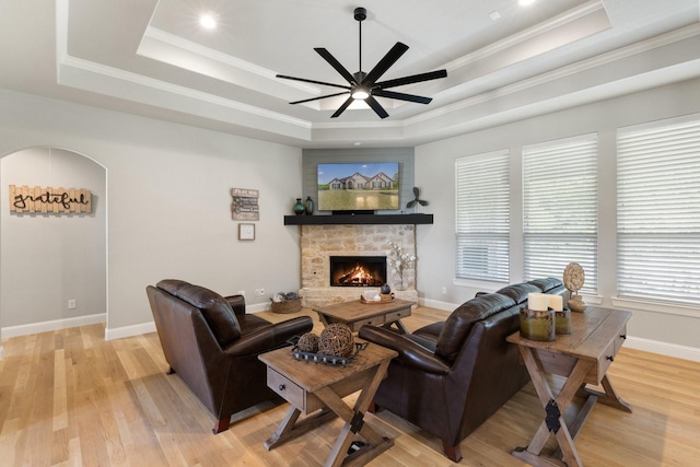 living room featuring a raised ceiling, a stone fireplace, a ceiling fan, ornamental molding, and light wood-type flooring