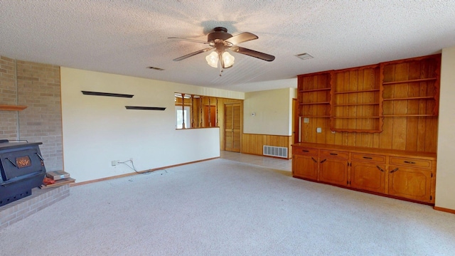 unfurnished living room with a wood stove, a ceiling fan, visible vents, a textured ceiling, and light colored carpet