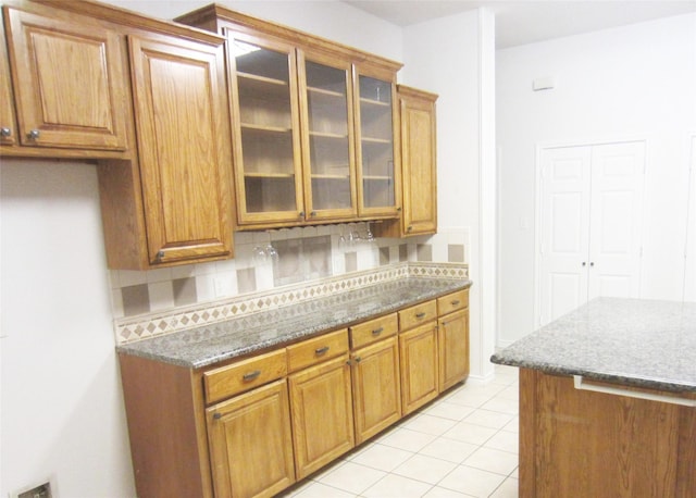 kitchen featuring brown cabinets, light tile patterned flooring, glass insert cabinets, stone counters, and tasteful backsplash
