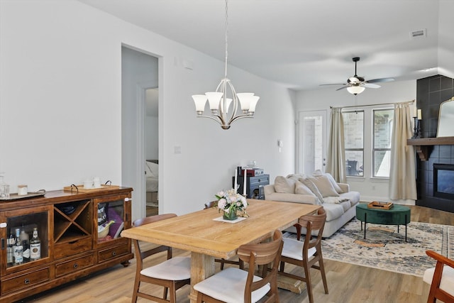 dining area featuring visible vents, light wood-type flooring, a tile fireplace, and ceiling fan with notable chandelier