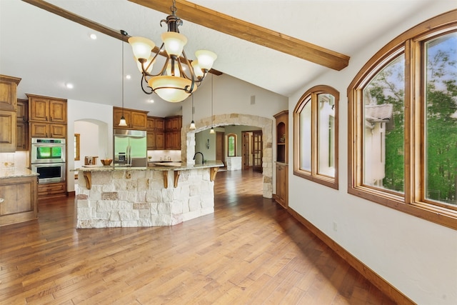 kitchen with appliances with stainless steel finishes, brown cabinetry, arched walkways, and beam ceiling