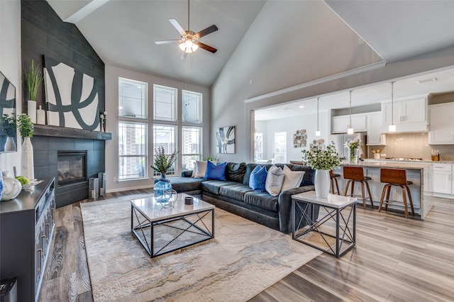 living area featuring a tiled fireplace, high vaulted ceiling, ceiling fan, and light wood-style flooring