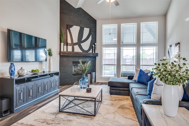 living area with a ceiling fan, high vaulted ceiling, a tiled fireplace, and wood finished floors
