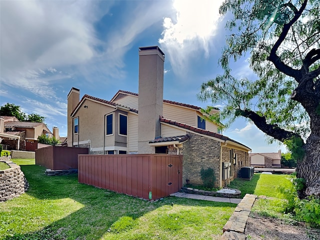 rear view of house featuring central air condition unit, a chimney, a lawn, stone siding, and fence
