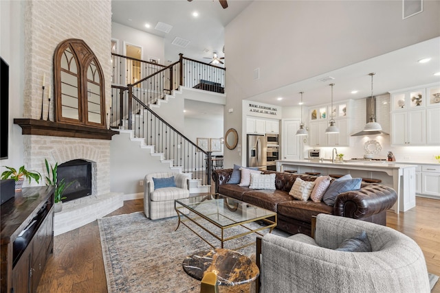 living area with a ceiling fan, stairs, a high ceiling, a brick fireplace, and dark wood-type flooring