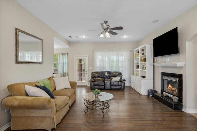 living area with a tile fireplace, a wealth of natural light, a ceiling fan, and dark wood-style flooring