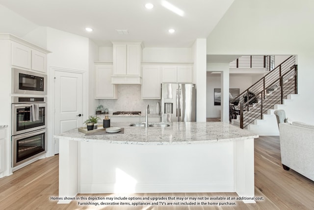 kitchen featuring a sink, an island with sink, stainless steel appliances, light wood-style flooring, and light stone countertops