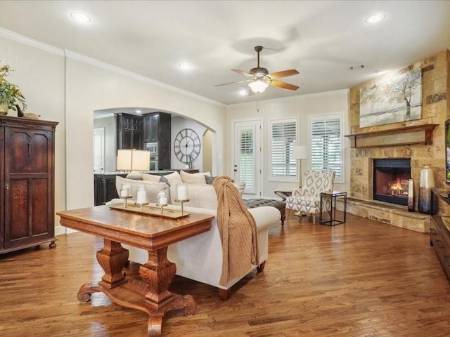 living room featuring a stone fireplace, wood finished floors, arched walkways, crown molding, and ceiling fan