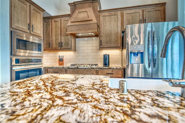 1023 Clydeview Rd, Forney, TX 75126 - kitchen with stainless steel appliances, tasteful backsplash, light stone counters, brown cabinetry, and premium range hood